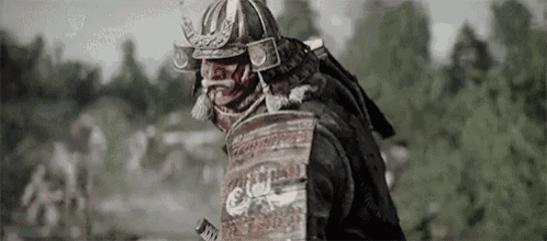 a samurai wearing armor and a helmet is standing in a field with trees in the background .