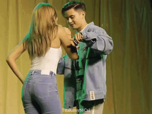 a man and a woman are dancing in front of a green curtain with titakimynical written on the bottom right corner
