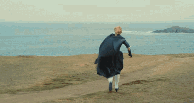 a woman in a black dress is running on a dirt path near the ocean