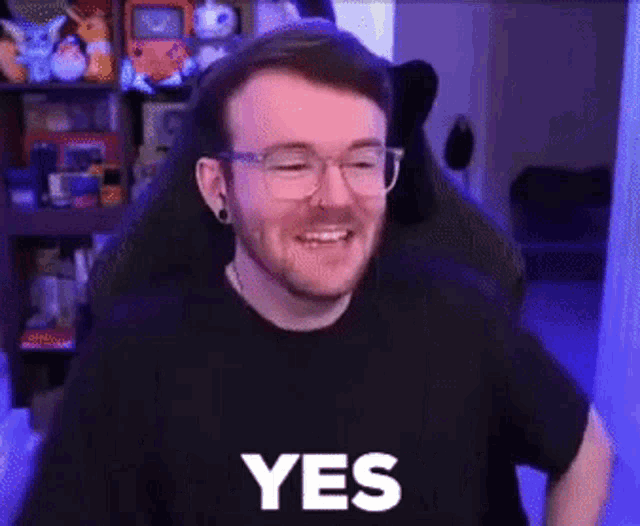 a man wearing glasses and a black shirt is sitting in a chair and smiling while saying yes .