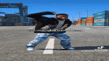 a man in a black jacket and camo pants is dancing on a street with containers in the background