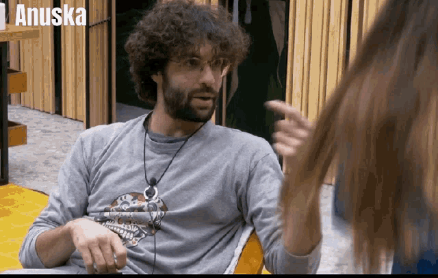 a man with a beard and glasses is talking to a woman with the name anuska behind him