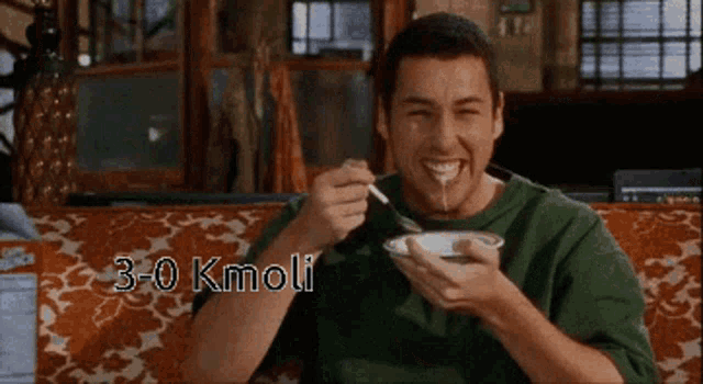 a man is sitting on a couch eating a bowl of food with the words 3-0 kmoli written below him