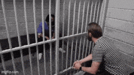 a man in a striped shirt is sitting in a jail cell talking to a man behind bars .