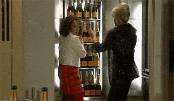 two women are standing in front of a fridge full of champagne bottles .