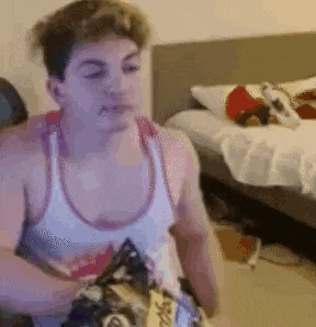 a man in a white tank top is sitting in front of a bed eating chips .
