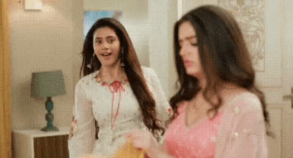 two women are standing next to each other in a living room . one of the women is wearing a pink dress .