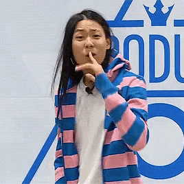 a woman wearing a pink and blue striped jacket holds her finger to her lips