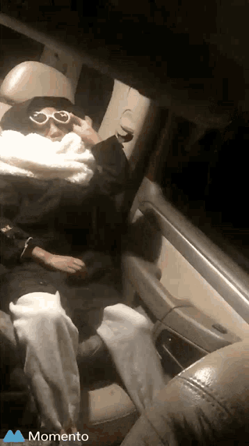 a person wearing sunglasses is sitting in the back seat of a car with a momento logo on the bottom right
