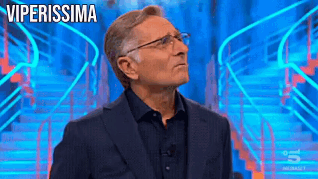 a man in a suit and glasses stands in front of a blue staircase with the words viperissima written on it