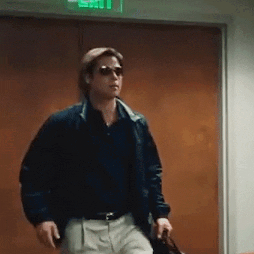 a man wearing sunglasses and a jacket is walking through a doorway holding a briefcase .