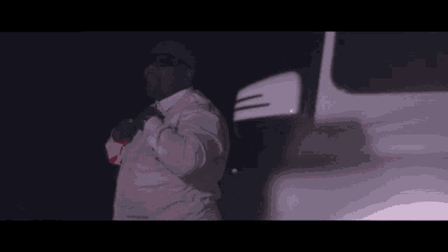 a man in a white shirt and sunglasses is standing next to a white car in the dark .