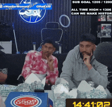 two men are sitting at a table with boxes of pizza and a clock that reads 14:21:02