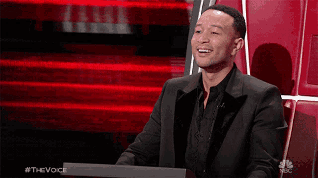 a man in a suit is sitting in front of a red wall with the words " the voice " on it