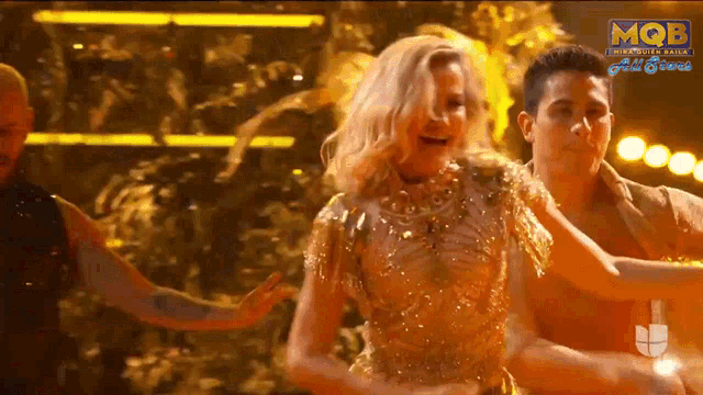 a woman in a gold dress is dancing with two men on a stage with mqb written on the bottom right