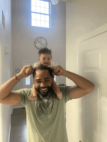 a man holds a baby on his shoulders in front of a clock that says xii on it