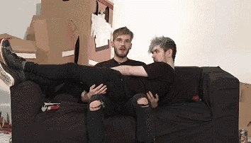 two men are sitting on a black couch and one of them is laying on the other 's lap