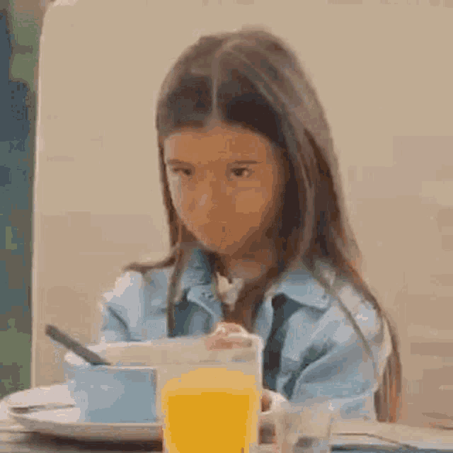a little girl is sitting at a table with a cup of orange juice and a bowl of cereal .