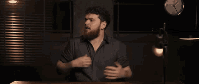 a man with a beard adjusts his shirt in front of a clock that shows the time as 7:15