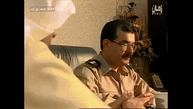 a man with glasses and a mustache is talking to another man with arabic writing on the screen