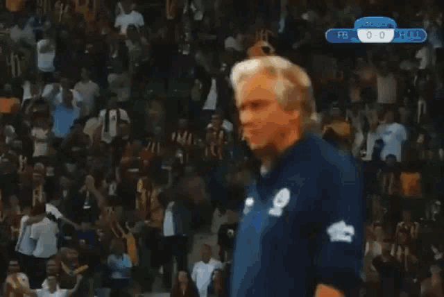 a man in a blue shirt stands in front of a crowd at a soccer game with the score 0-0