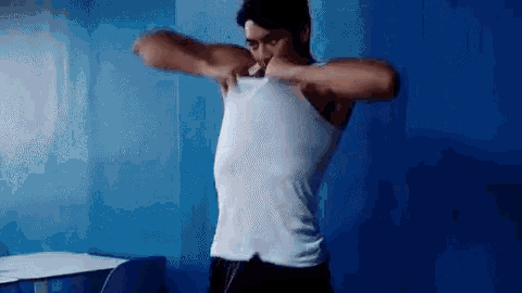 a man in a white tank top is standing in front of a blue wall and taking off his shirt .