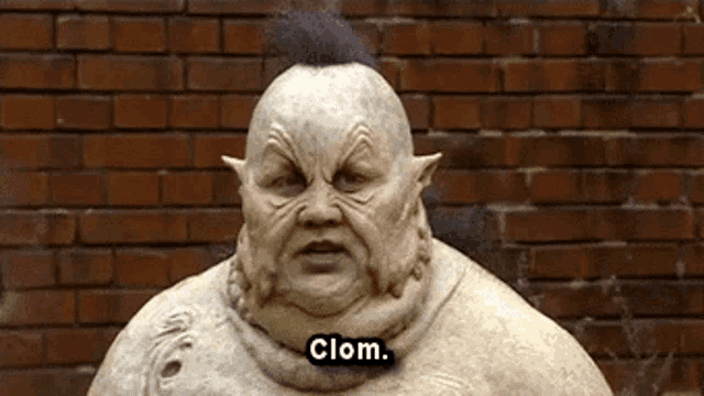 a statue of a clown is standing in front of a brick wall and saying clom .