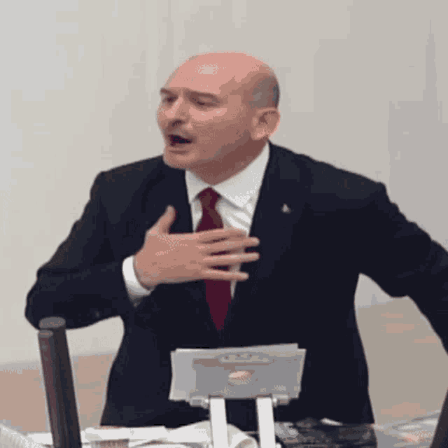 a man in a suit and tie is sitting at a desk with his hand on his chest