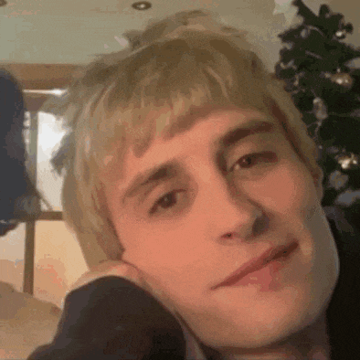 a young man with blonde hair is sitting in front of a christmas tree and smiling .