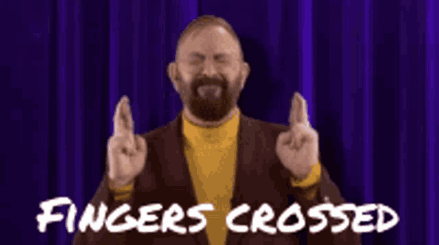 a man in a suit and yellow turtleneck is crossing his fingers in front of a blue curtain .