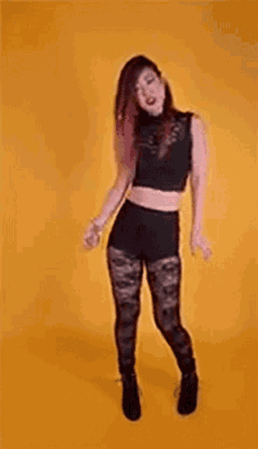 a woman is dancing in front of a yellow background . she is wearing a black crop top and black leggings .