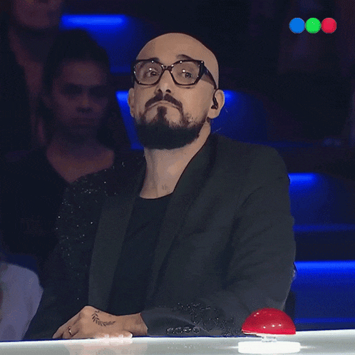 a bald man with glasses and a beard sits at a table with a red button in front of him
