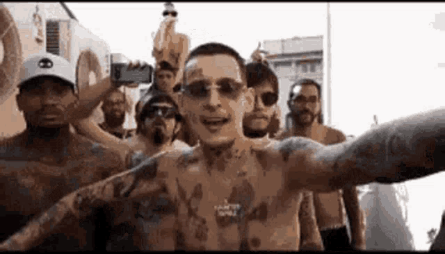 a group of men are standing around a shirtless man wearing sunglasses and a hat .