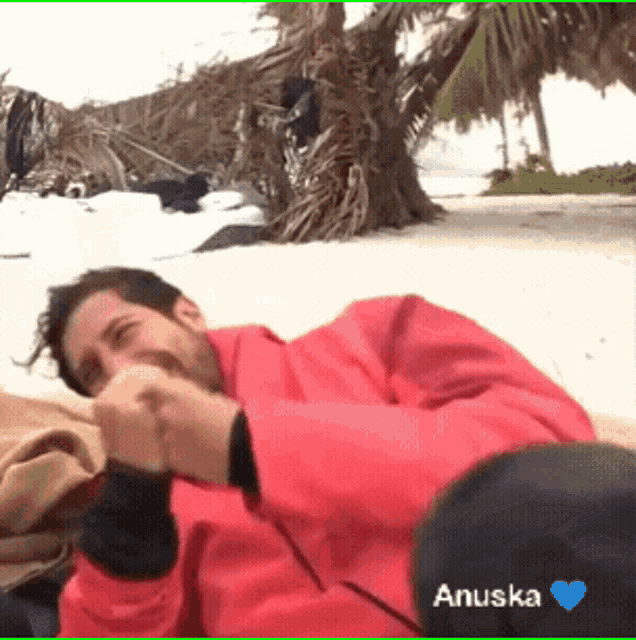 a man in a red jacket is laying on a beach next to a tree and the name anuska