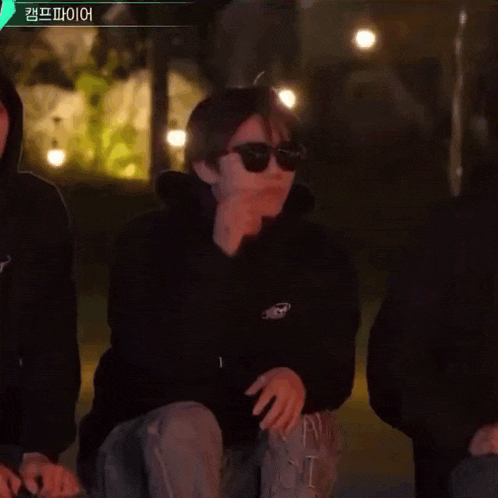 a man wearing sunglasses and a black hoodie is kneeling down in front of a group of people .