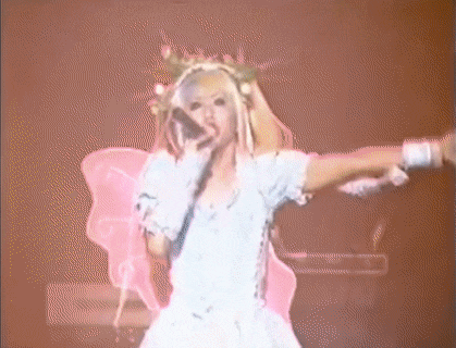 a woman in a white dress and pink wings is singing into a microphone on a stage .