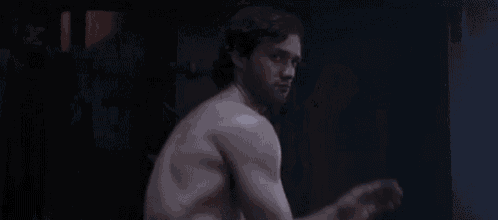 a shirtless man is standing in a dark room with his hands up .