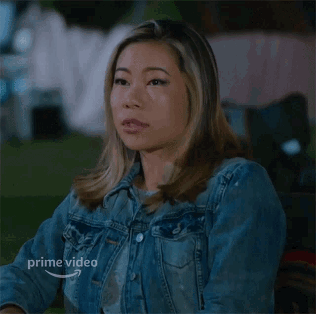 a woman in a denim jacket is sitting in a chair with a prime video logo on the bottom right
