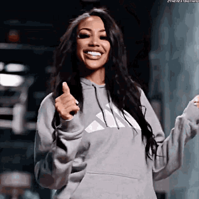 a woman wearing a gray adidas hoodie is smiling and giving a thumbs up