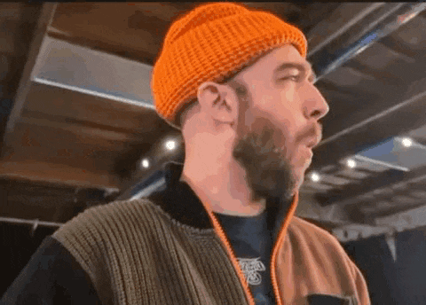 a man with a beard wearing an orange beanie and a brown jacket is standing in a room .