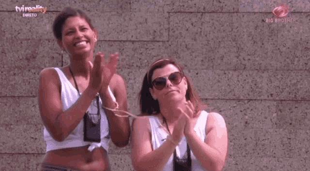 two women are clapping in front of a brick wall with a tvreality logo in the corner
