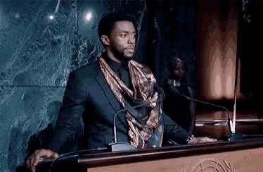 a man in a suit and scarf is standing at a podium with microphones .