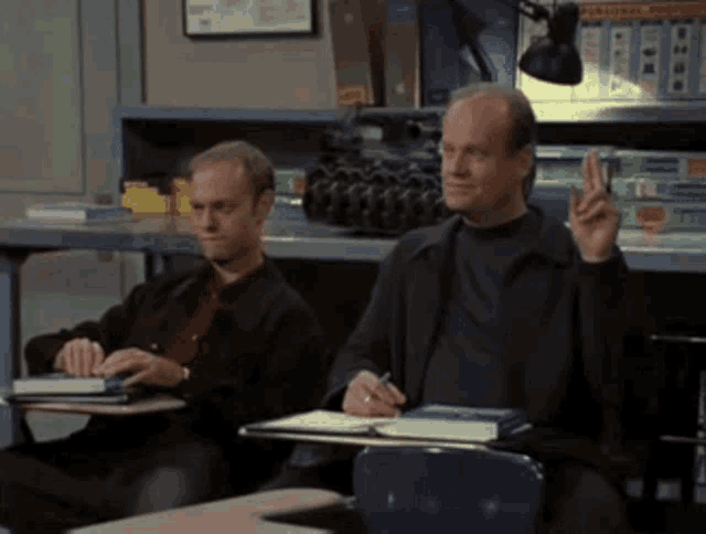 two men are sitting at desks in a classroom and one is pointing