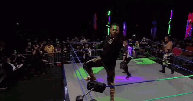 a wrestler is jumping in the air in a wrestling ring while a referee looks on .