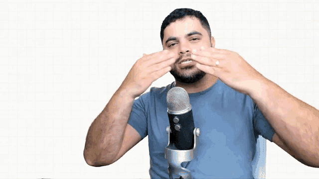 a man in a blue shirt stands in front of a microphone covering his mouth with his hands