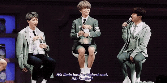 jimin has the highest seat and jm smiles while sitting on a chair