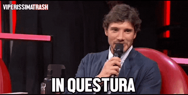 a man in a suit is holding a microphone and the word in questura is above him
