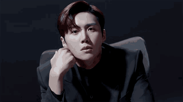 a man in a black suit is sitting in a chair with his hand on his chin