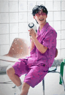 a man in a purple shirt and shorts is sitting on a chair holding a hair dryer and blowing bubbles .