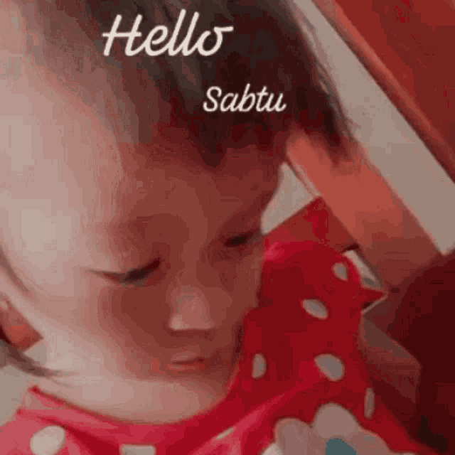 a baby in a red polka dot shirt is sitting on a chair and says hello sabtu .
